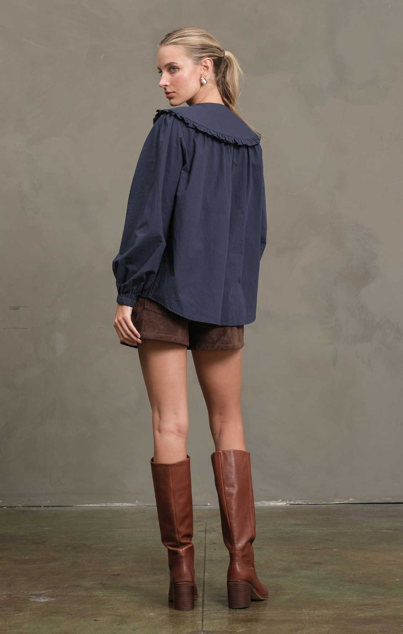 Woman wearing a navy blue blouse, brown shorts, and brown knee-high boots against a plain background