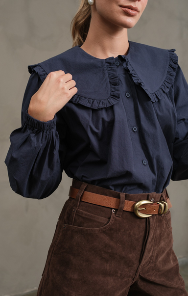 Person wearing a navy blue blouse with ruffled collar and brown pants against a neutral background