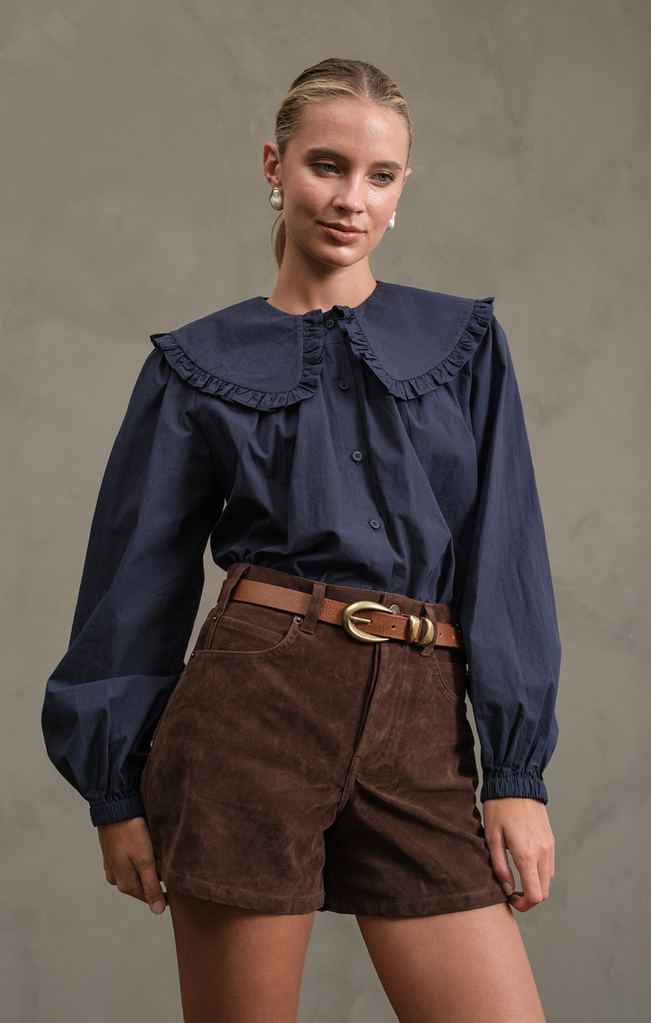 Woman wearing a navy blue blouse with ruffled collar and brown shorts against a gray background