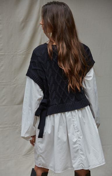 Person wearing a black knitted cardigan over a white dress against a plain background