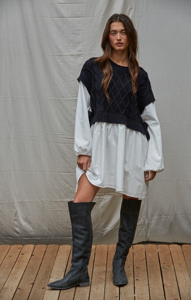 Woman wearing a white dress, black quilted vest, and knee-high black boots against a plain background.