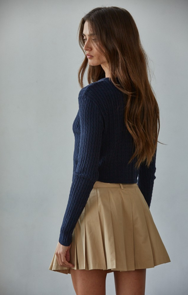 Woman wearing a navy sweater and beige pleated skirt against a plain background