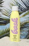 Yellow sunscreen spray bottle with purple logo against a palm leaf background