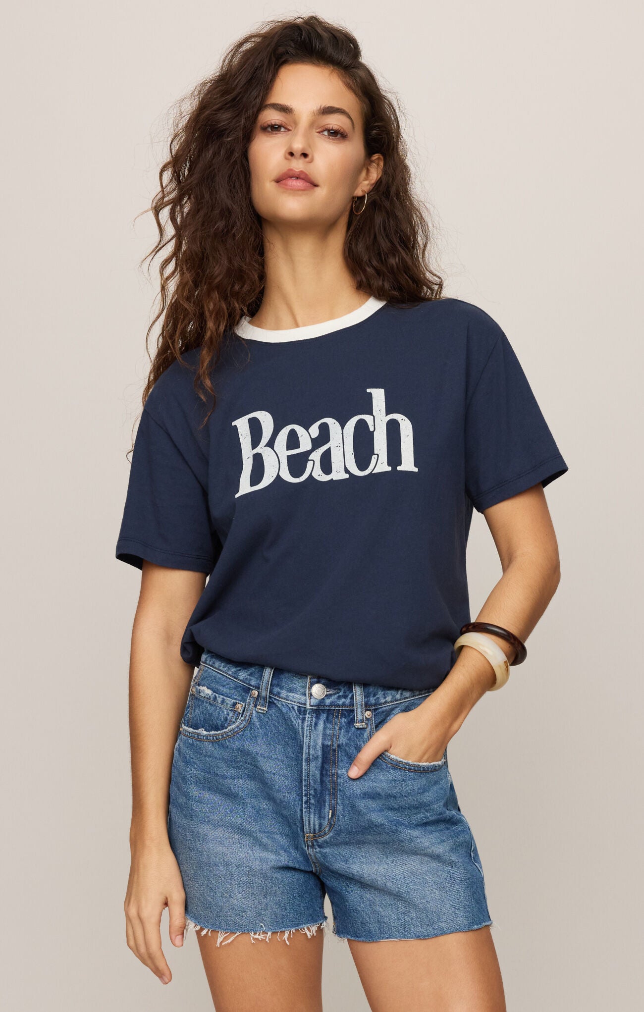Woman wearing a navy blue t-shirt with 'Beach' text and denim shorts on a beige background
