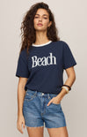 Woman wearing a navy blue t-shirt with 'Beach' text and denim shorts on a beige background