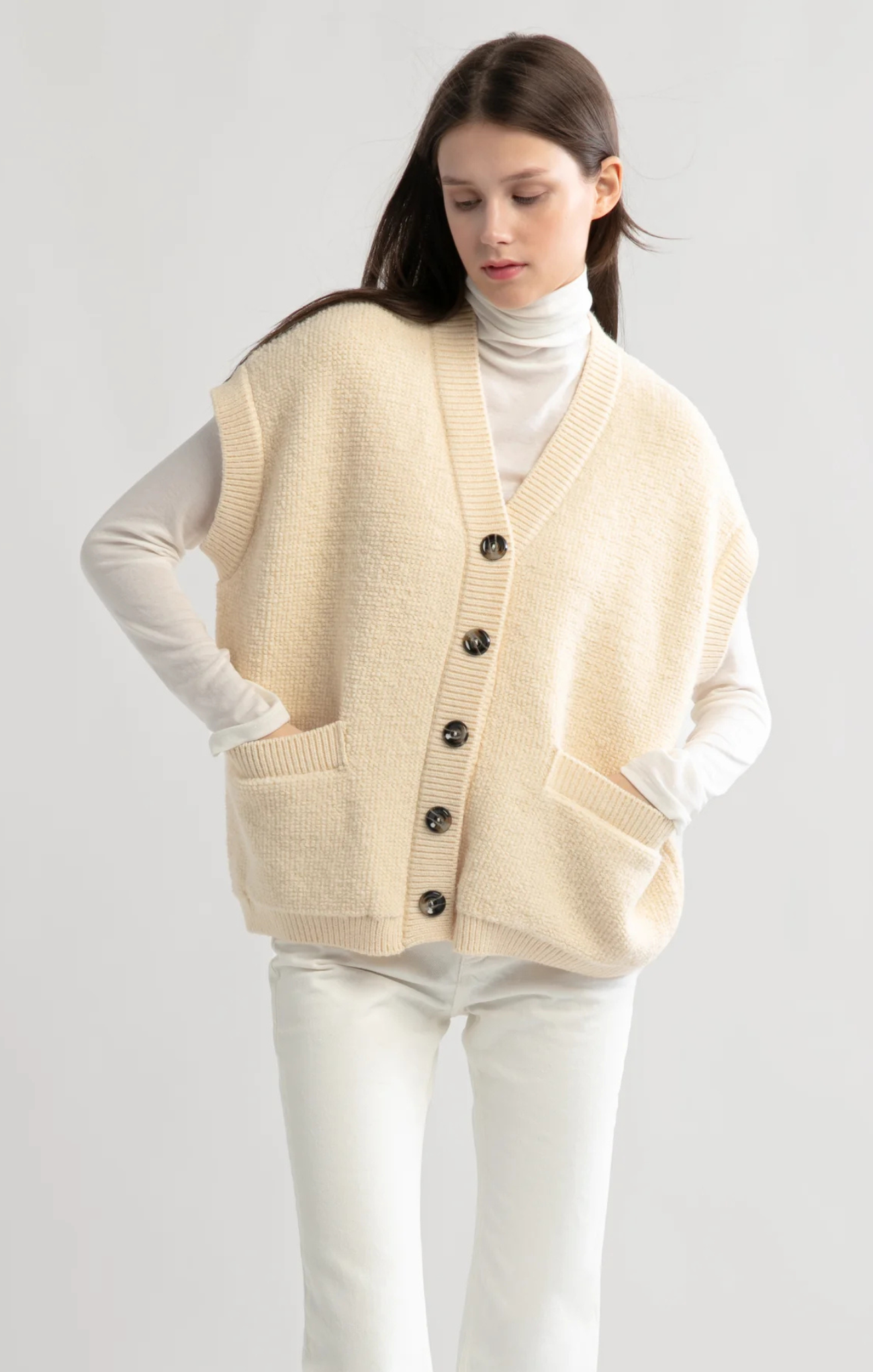 Woman wearing a beige cardigan over a white turtleneck and pants on a light gray background