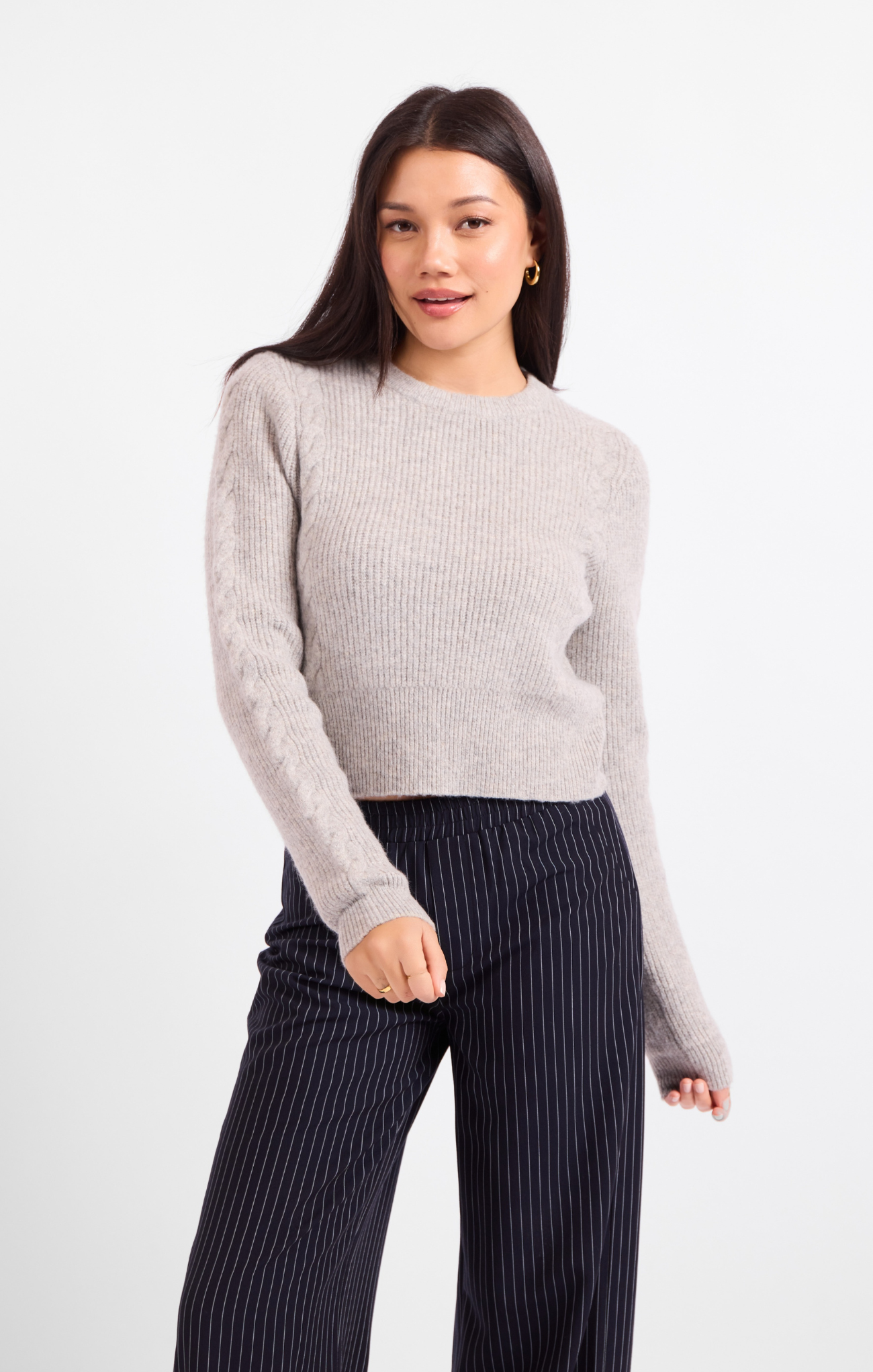 A woman modeling a light gray ribbed knit sweater paired with dark navy pinstriped trousers, shot against a clean white studio background.