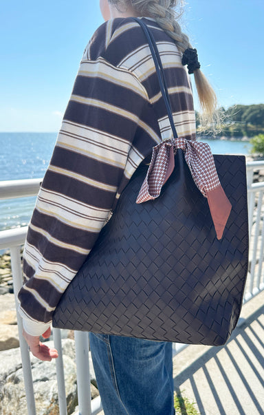 Person holding a black woven handbag with a checkered ribbon by a waterfront