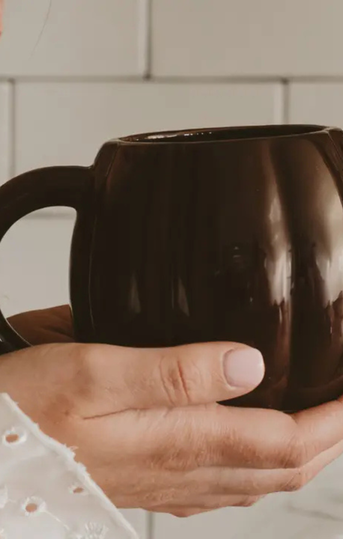 A close-up of a person's hands holding a glossy dark brown ceramic mug against a light tiled background.