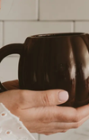 A close-up of a person's hands holding a glossy dark brown ceramic mug against a light tiled background.