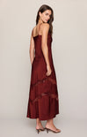 A woman modeling a sleeveless, floor-length burgundy slip dress with sheer lace trim details near the lower skirt, shown from a three-quarters back view against a neutral studio background.