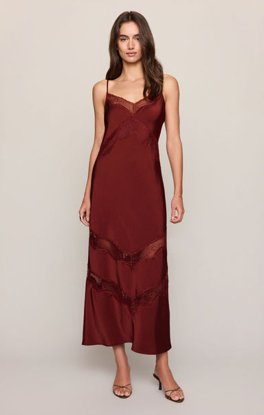 A woman modeling a long burgundy satin slip dress with lace trim and spaghetti straps against a plain light gray studio background.