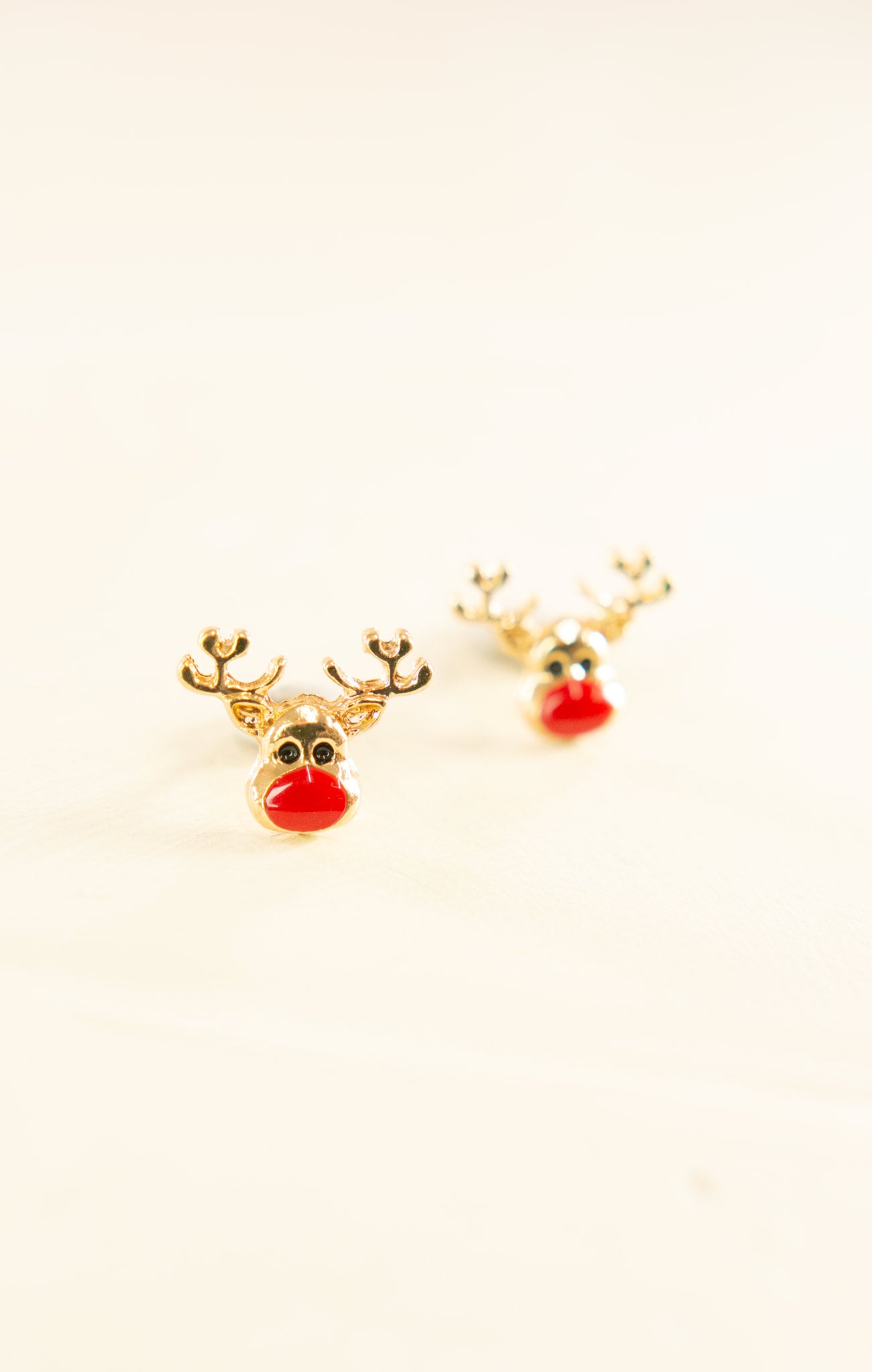 Gold reindeer-shaped earrings with red noses on a light beige background