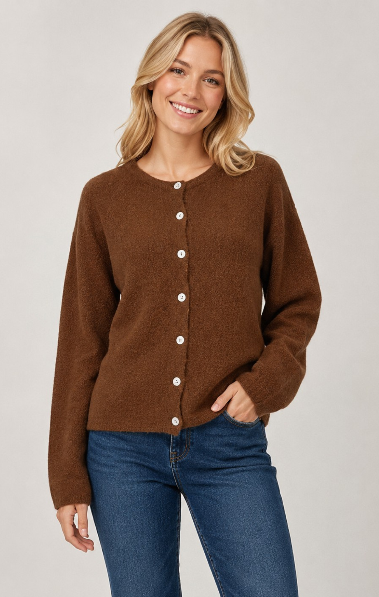 A woman wearing a brown button-front cardigan sweater and blue jeans, standing against a light gray studio background.