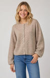 A woman wearing a beige button-front knit cardigan with balloon sleeves and blue jeans, standing against a light gray studio background.