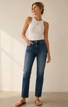 A woman modeling a pair of high-waisted, straight-leg blue jeans with a white ribbed tank top and brown sandals, standing against a warm beige wall.