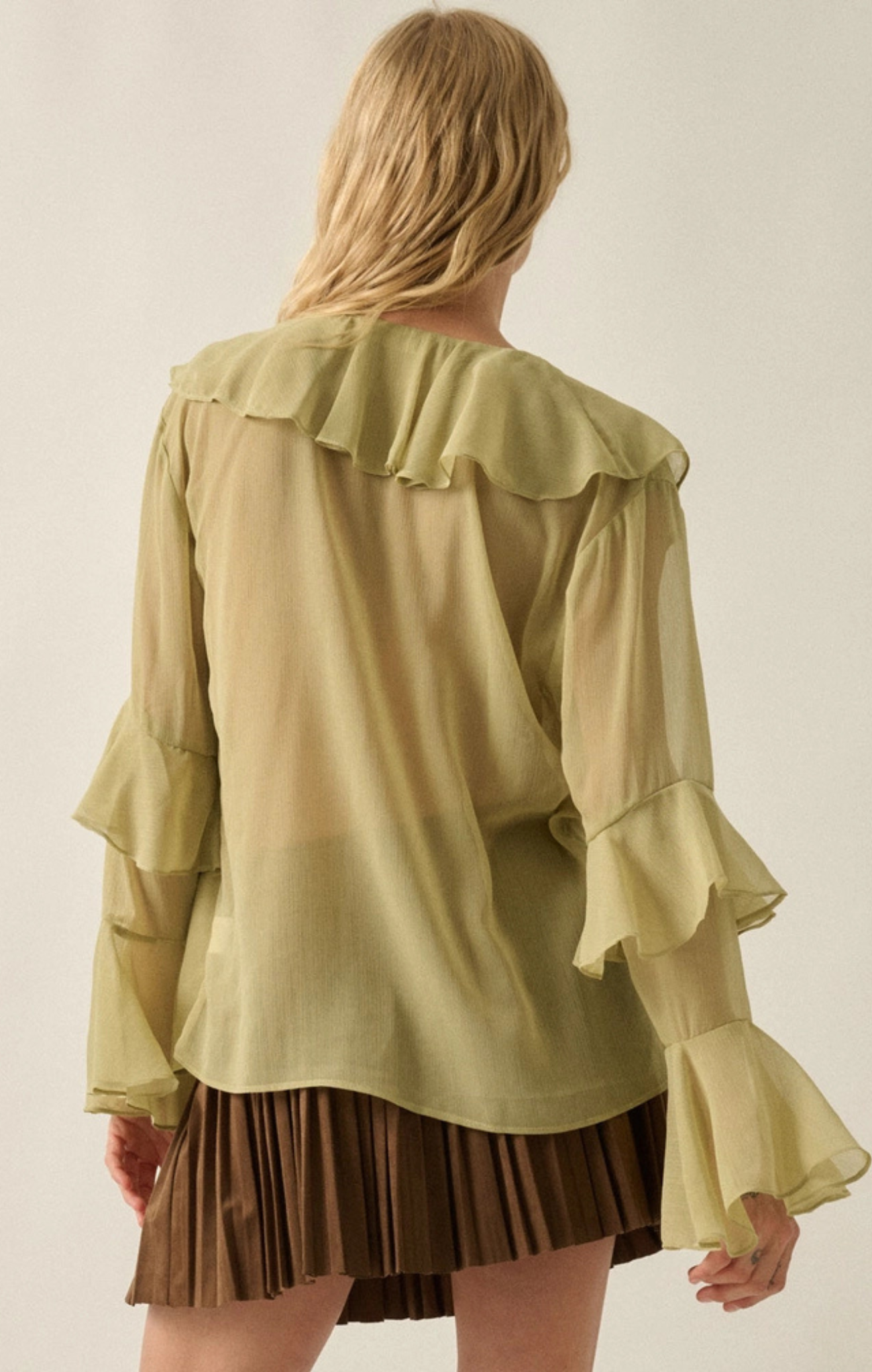 A woman shown from the back wearing a sheer olive green blouse with layered ruffles and long flared sleeves, paired with a brown pleated skirt, against a plain light beige studio background.