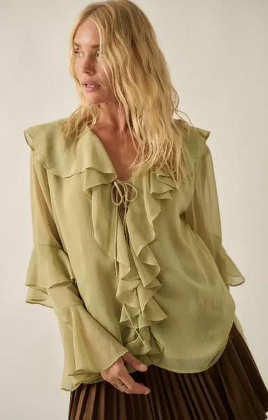 A woman wearing a sage green ruffled blouse with long sleeves and a tie at the neckline, paired with a brown pleated skirt, against a plain light beige background.