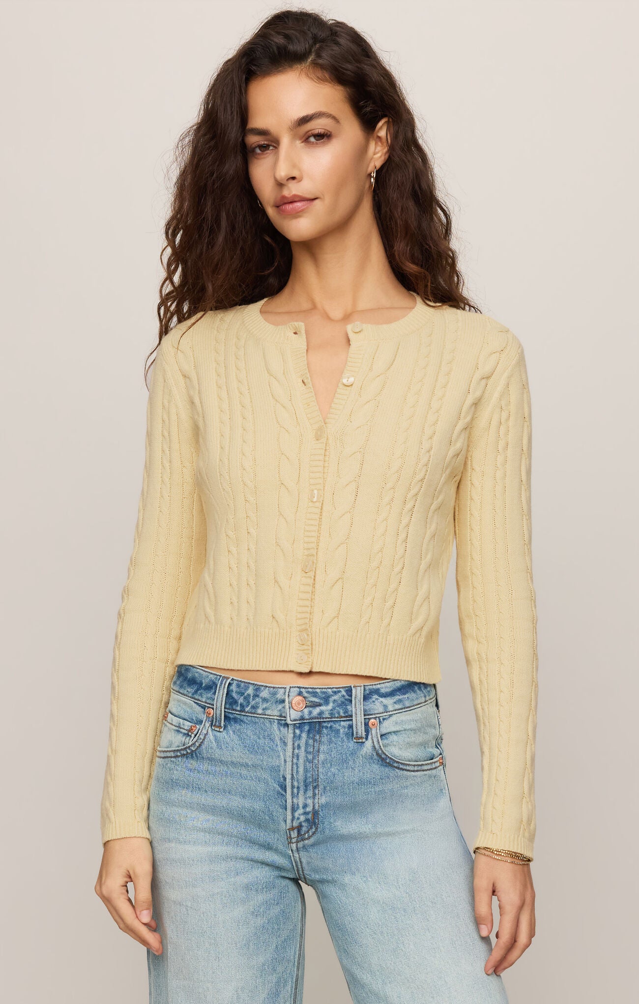 Woman wearing a light yellow cable knit cardigan and blue jeans against a plain background