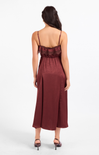 A woman viewed from the back wearing a sleeveless midi dress in a deep burgundy or maroon shade with thin straps and a ruffled neckline, paired with dark heeled sandals against a plain light gray studio background.