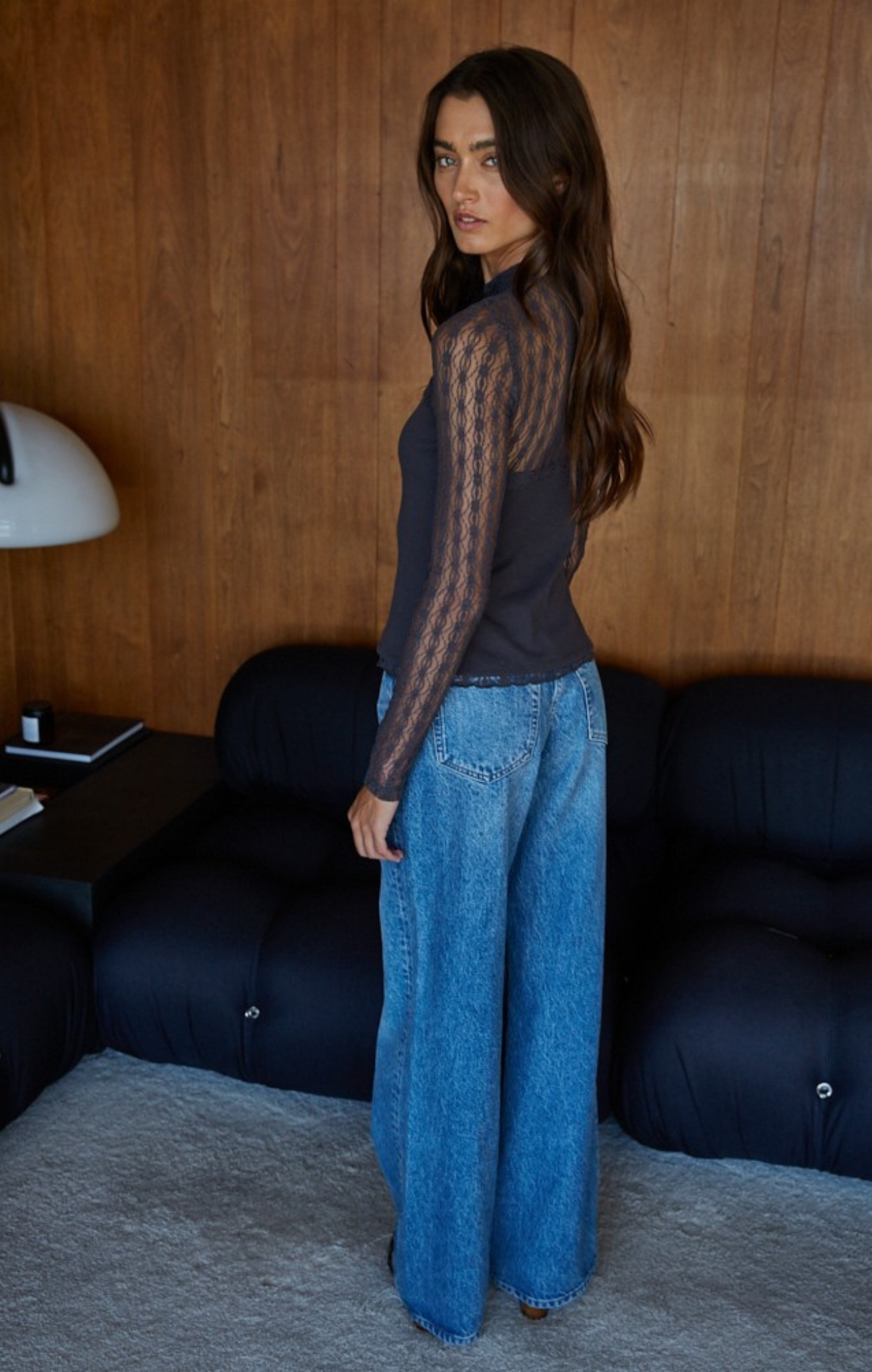 Woman wearing a black top with sheer sleeves and blue jeans in a room with wooden walls and a dark sofa.
