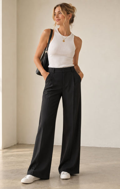 A woman stands in a minimalist studio wearing black wide-leg trousers, a white sleeveless top, white sneakers, and carrying a black shoulder bag.