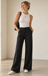 A woman stands in a minimalist studio wearing black wide-leg trousers, a white sleeveless top, white sneakers, and carrying a black shoulder bag.
