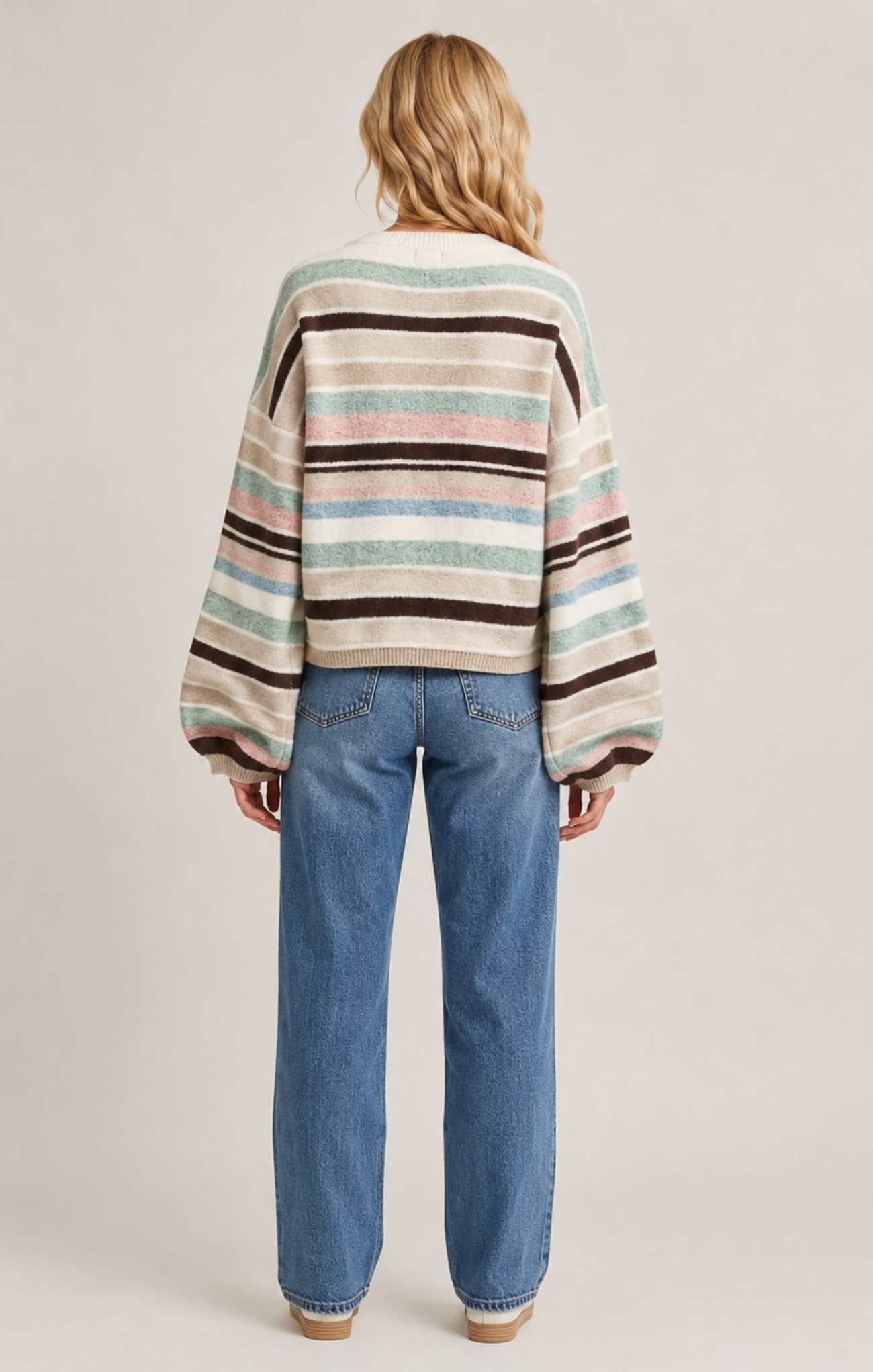 A woman viewed from the back wearing a loose striped knit sweater with beige, brown, mint green, and pink horizontal stripes, paired with blue jeans, against a plain light beige studio background.