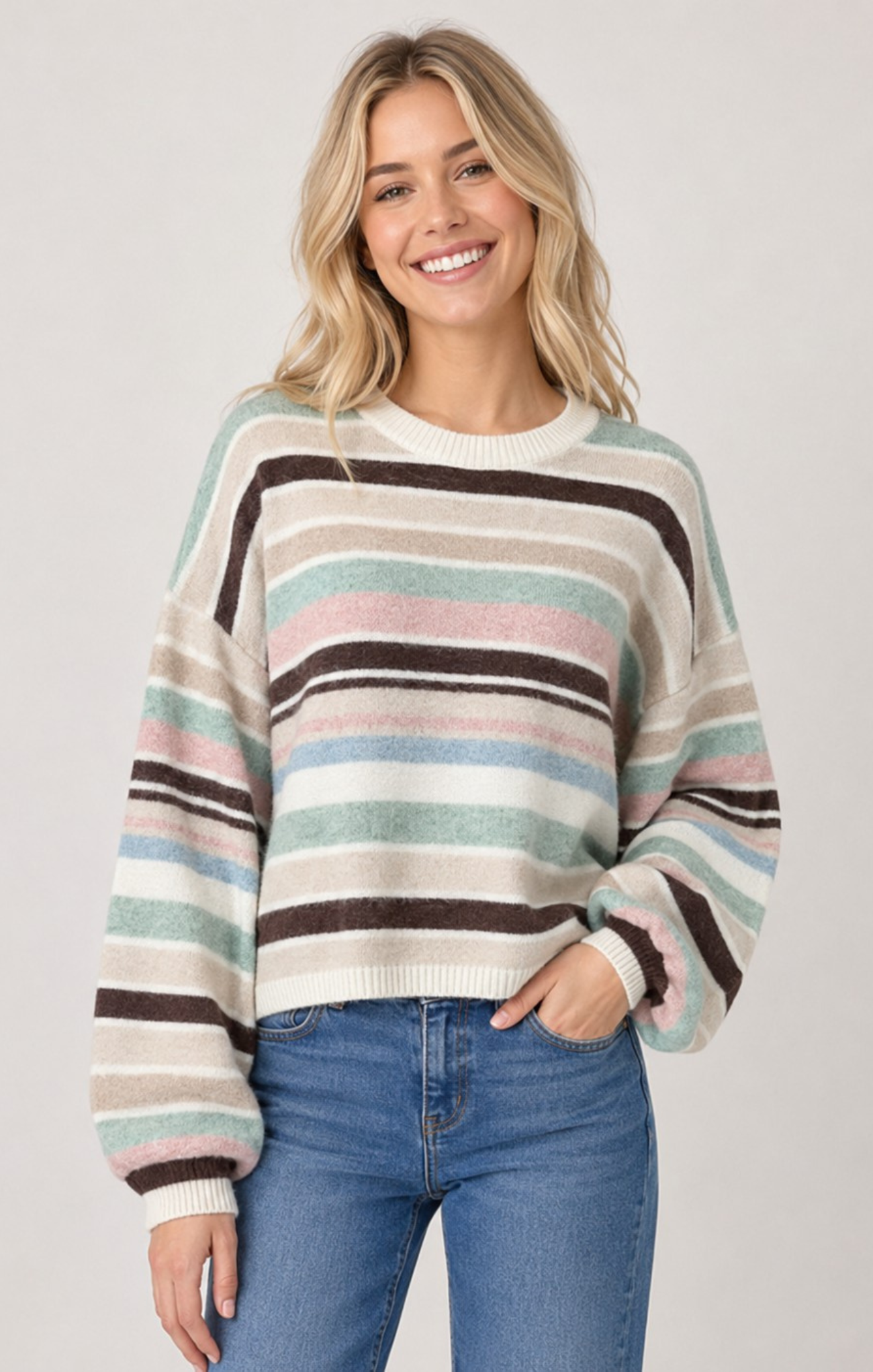 A woman wearing a long-sleeve crewneck sweater with horizontal stripes in cream, brown, light blue, and pink, paired with blue jeans, against a light gray studio background.