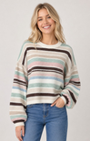 A woman wearing a long-sleeve crewneck sweater with horizontal stripes in cream, brown, light blue, and pink, paired with blue jeans, against a light gray studio background.