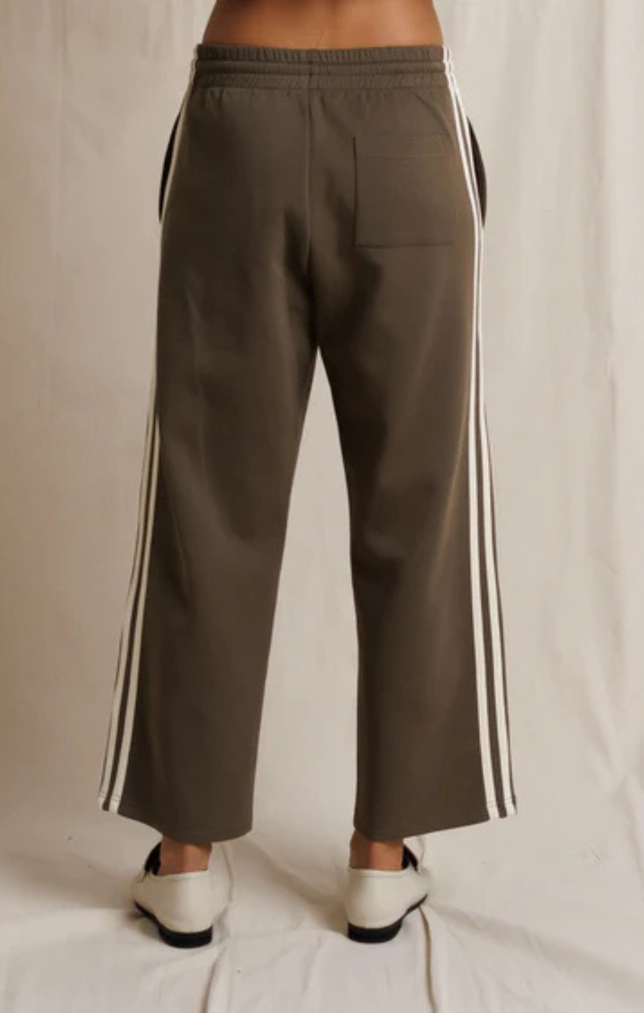 A person shown from the back wearing wide-leg cropped pants in a muted olive-brown color with white side stripes, paired with white shoes, against a light fabric backdrop.