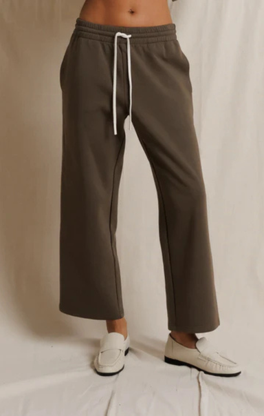 A person wearing wide-leg cropped sweatpants in a muted taupe-brown color with a white drawstring, paired with white loafers, standing against a draped off-white fabric backdrop.