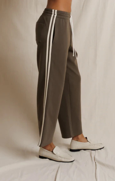 A person wearing wide-leg taupe-brown pants with white side stripes and white loafers, standing against a draped off-white fabric backdrop.