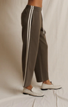 A person wearing wide-leg taupe-brown pants with white side stripes and white loafers, standing against a draped off-white fabric backdrop.