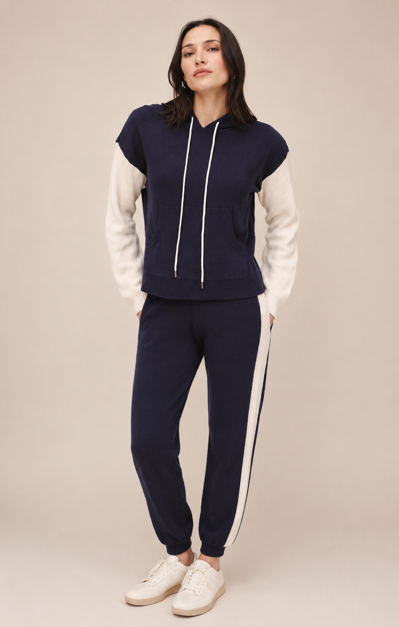 A woman modeling a navy blue and white color-blocked tracksuit with a cropped hoodie top and matching jogger pants, paired with white sneakers, against a solid beige studio background.