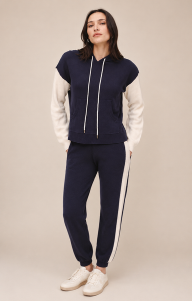 A woman modeling a navy blue and white color-blocked tracksuit with a cropped hoodie top and matching jogger pants, paired with white sneakers, against a solid beige studio background.