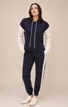 A woman modeling a navy blue and white color-blocked tracksuit with a cropped hoodie top and matching jogger pants, paired with white sneakers, against a solid beige studio background.