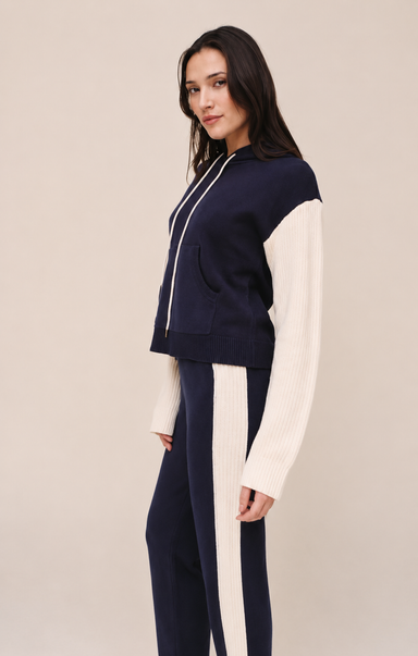 A woman modeling a navy blue and off-white color-blocked zip-up hoodie and matching sweatpants against a solid beige studio background.