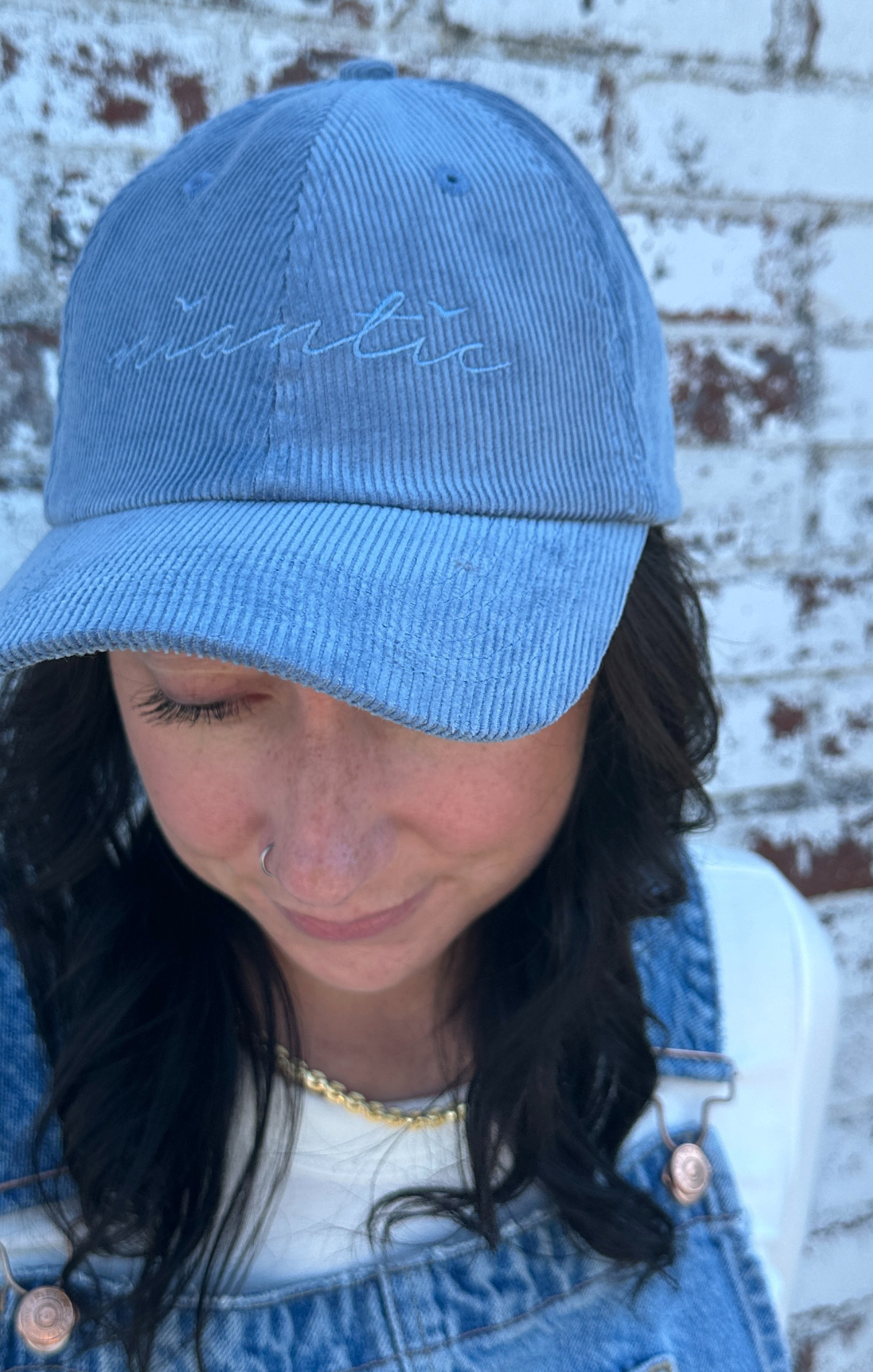 Person wearing a blue cap with 'nana' embroidered on it against a brick wall.