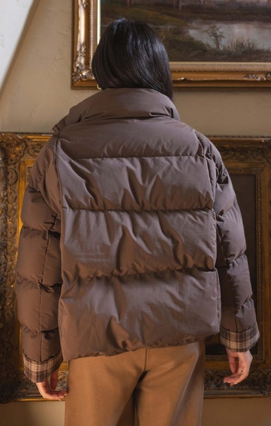A person viewed from behind wearing a brown puffer jacket with horizontal quilting, paired with tan pants, standing indoors in front of ornate framed artwork.