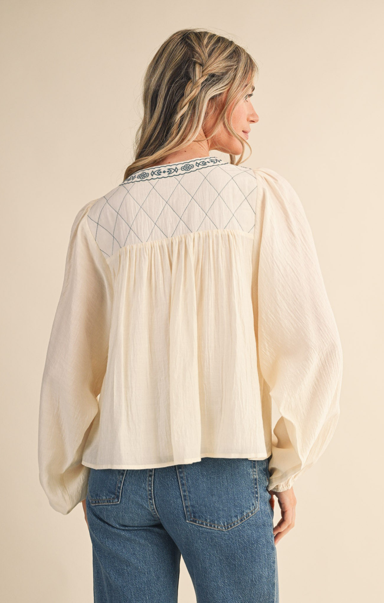Woman wearing a beige blouse with embroidered details and blue jeans on a beige background
