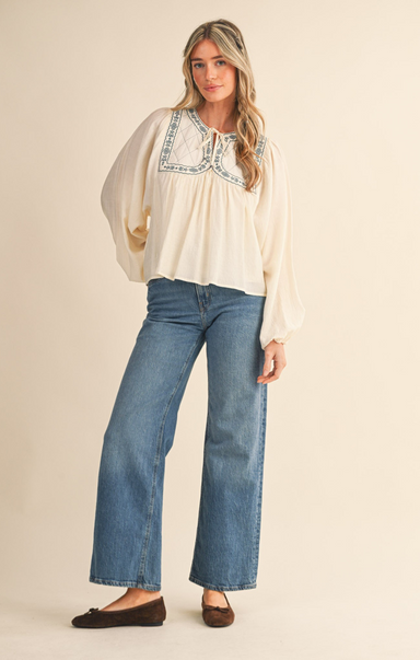 Woman wearing a white blouse with blue embroidery and blue jeans on a beige background