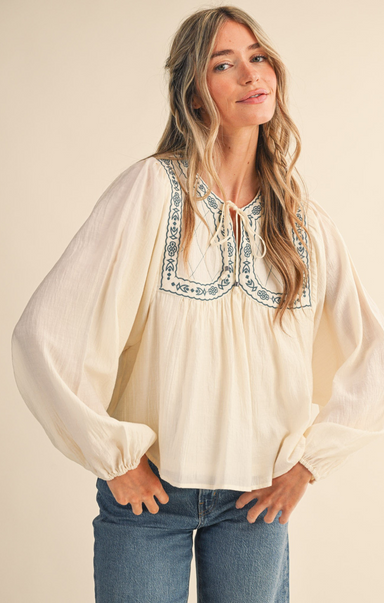 Woman wearing a cream blouse with blue embroidery and jeans on a beige background