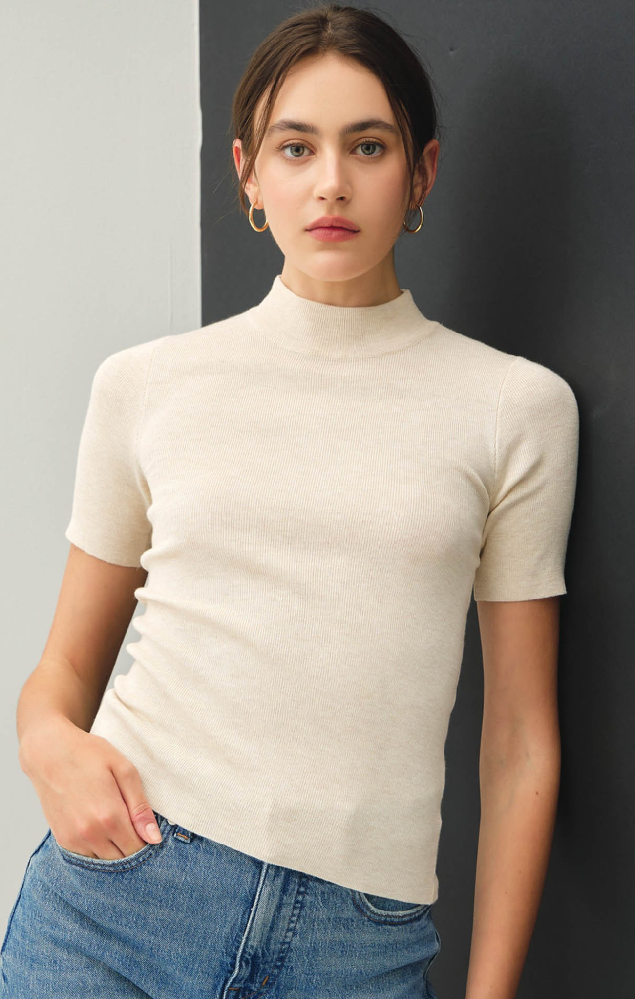Woman wearing a beige turtleneck sweater and blue jeans against a gray wall.
