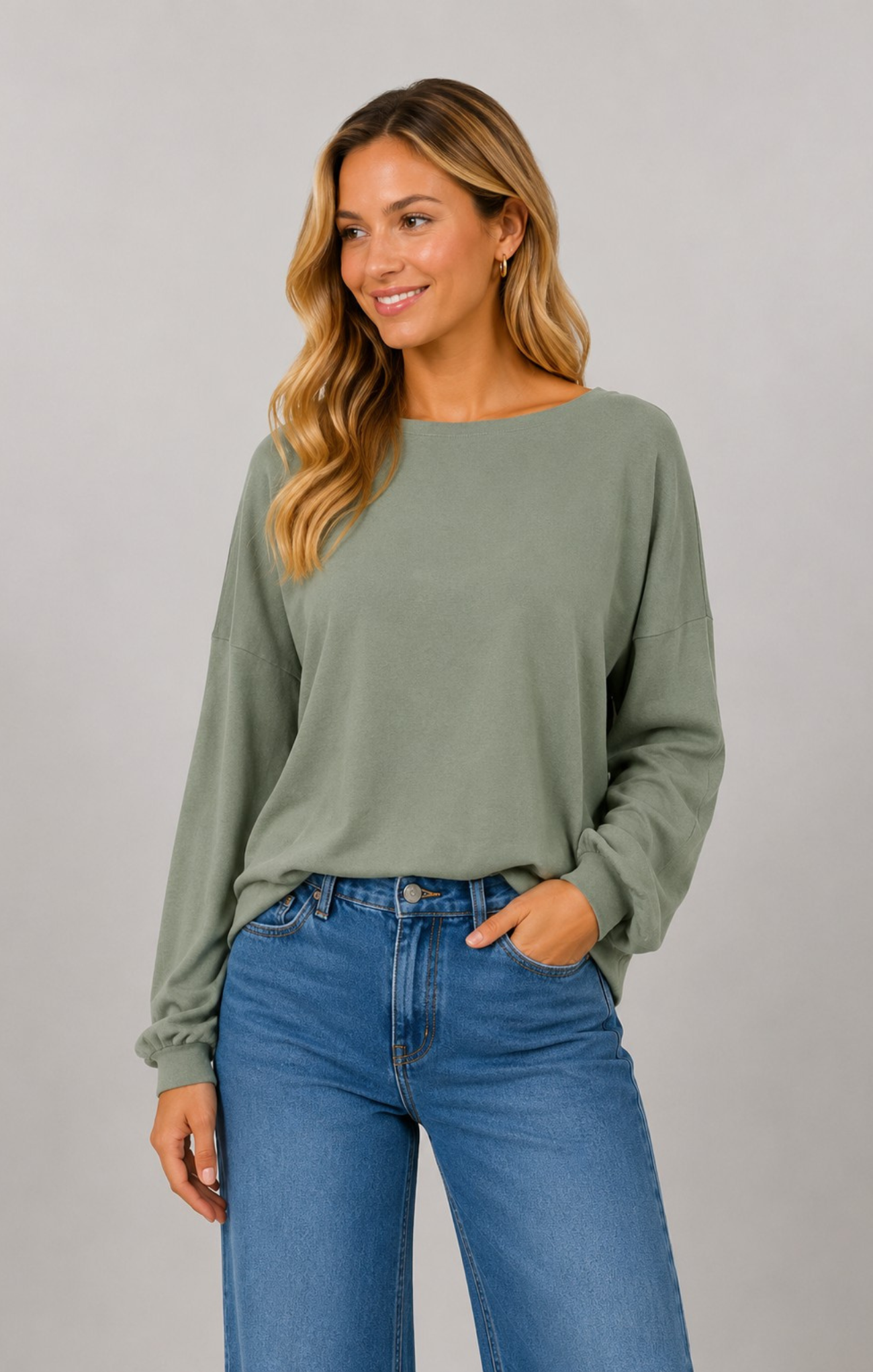 A woman wearing a sage green long-sleeve knit top and blue jeans, standing against a solid gray studio background.