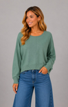 A woman wearing a sage green long-sleeve crewneck sweater and blue jeans, standing against a solid gray studio background.