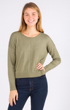 A woman modeling a long-sleeve crewneck knit sweater in sage green, paired with dark blue jeans, against a plain white studio background.