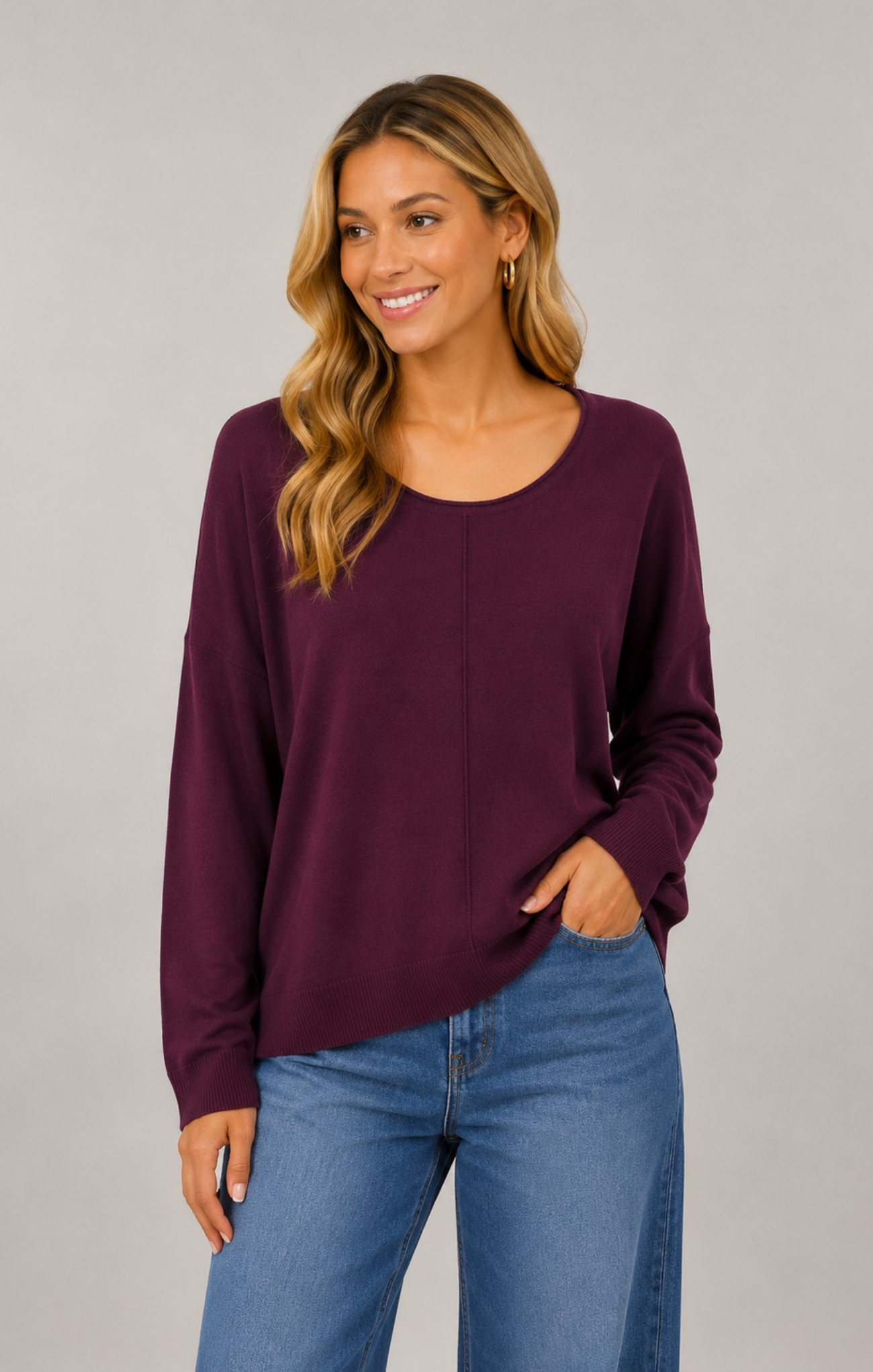 A woman wearing a long-sleeve scoop-neck knit top in deep plum purple, paired with blue jeans, standing against a solid light gray studio background.