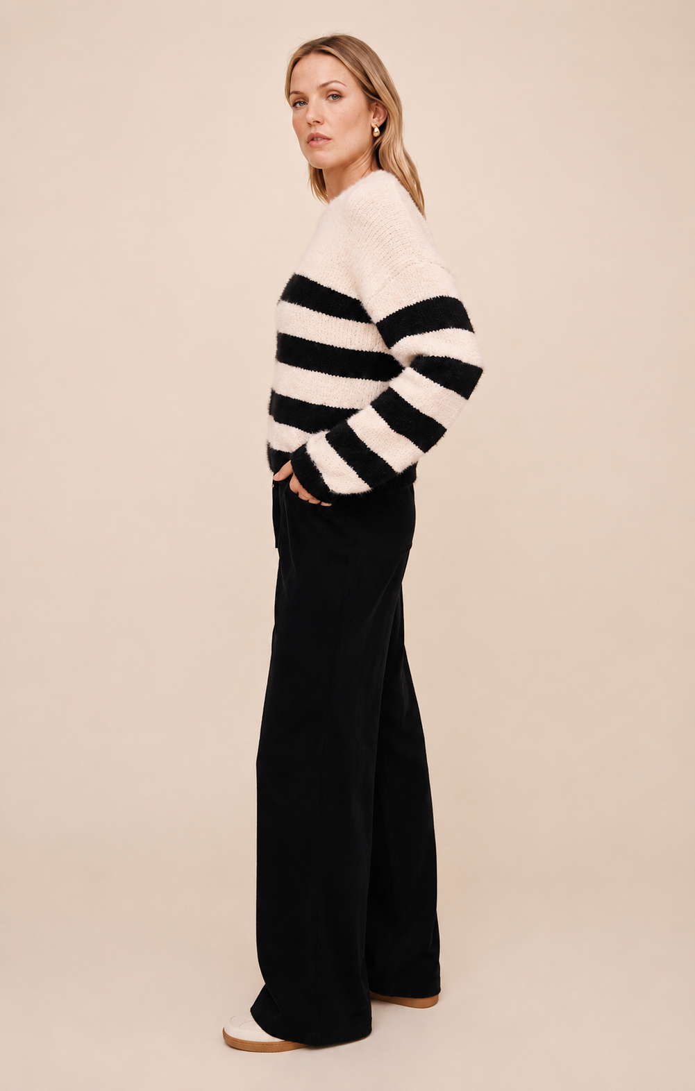 A woman stands in profile wearing a fuzzy knit sweater with wide black and off-white horizontal stripes, paired with black wide-leg pants, against a plain beige studio background.