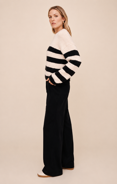 A woman stands in profile wearing a fuzzy knit sweater with wide black and off-white horizontal stripes, paired with black wide-leg pants, against a plain beige studio background.